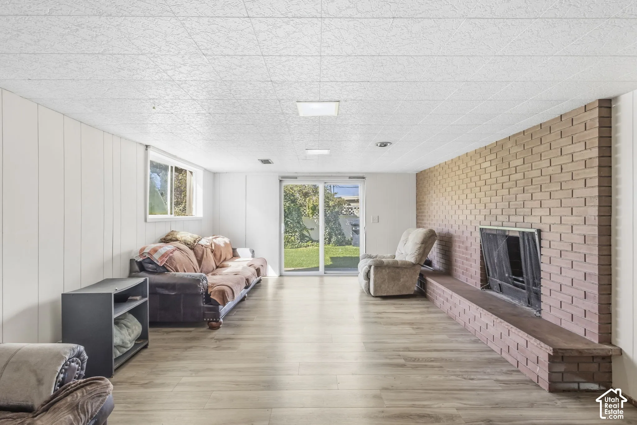 Wood-finish flooring, brick-faced fireplace, white paneled walls, and sliding glass door with independent access directly from private backyard
