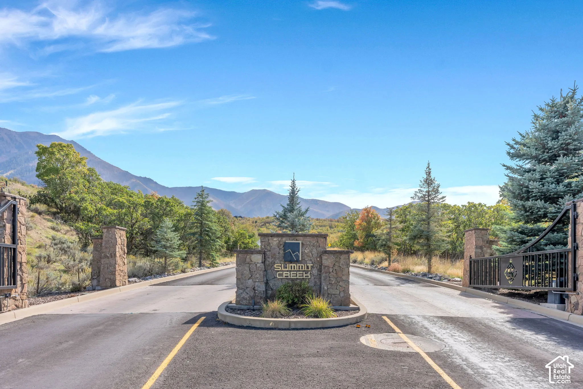 Stone-clad entry pillars with wrought iron gates, a central stone monument, and paved roadway with natural landscaping