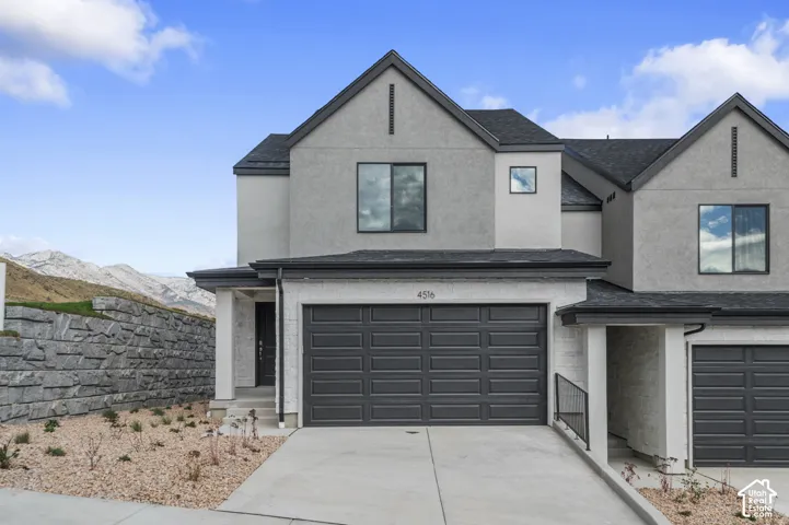 Stucco exterior with gabled roof lines, dark garage door, concrete driveway, and stone retaining wall