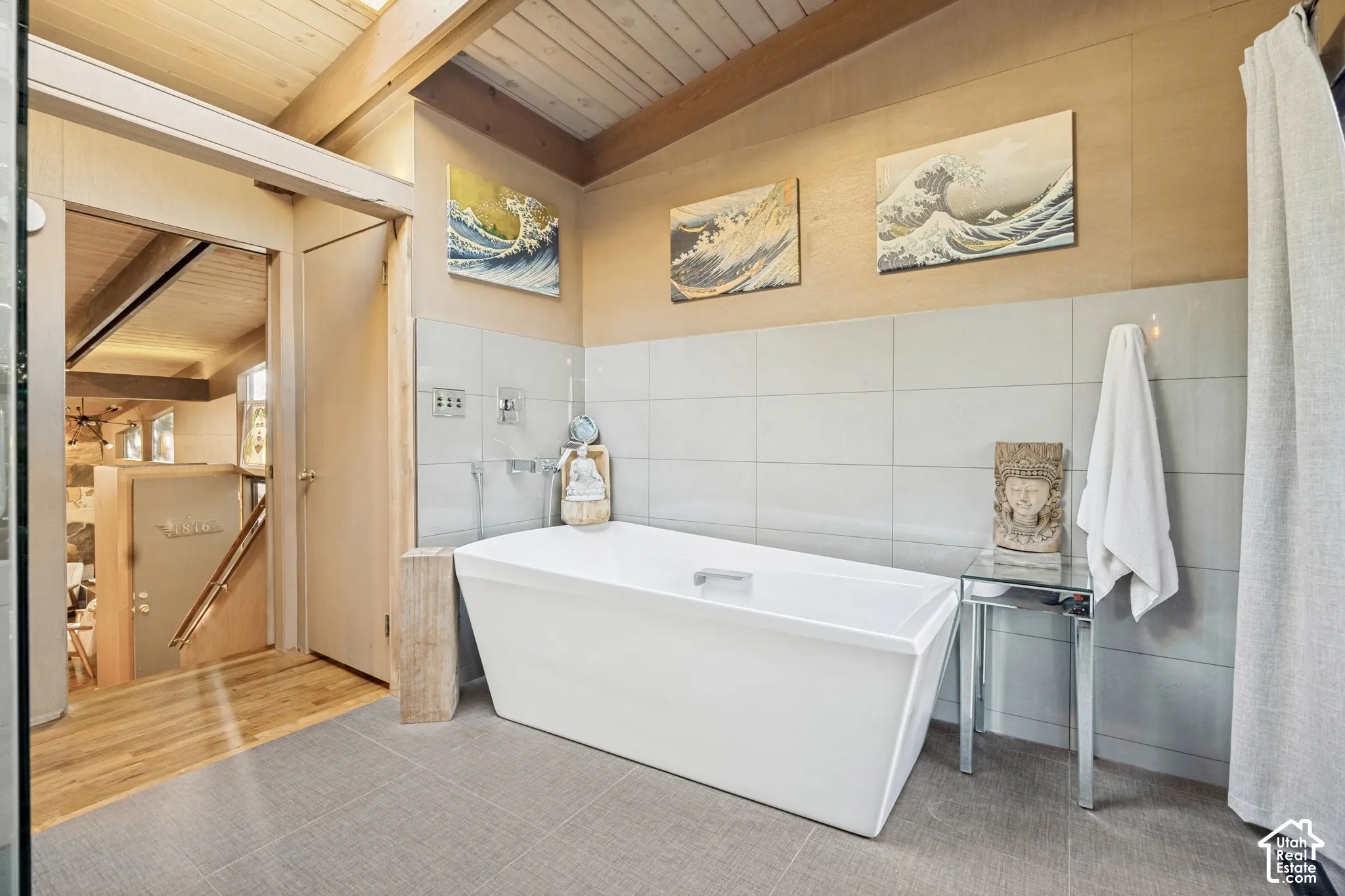 Freestanding soaking tub with polished chrome fixtures, light gray tile flooring, and wood-paneled ceiling with exposed beams
