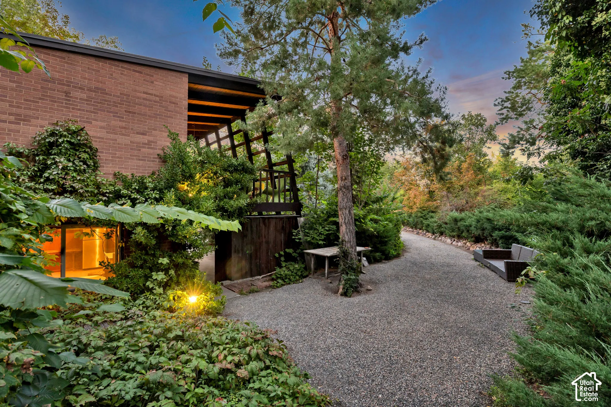 Brick exterior with a covered patio and trellis, complemented by extensive landscaping with mature trees and a gravel pathway