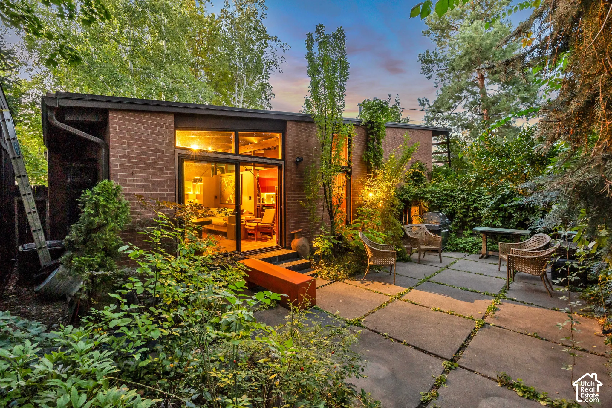 Brick exterior with expansive glass doors and windows, leading to a concrete paver patio surrounded by lush landscaping and mature trees