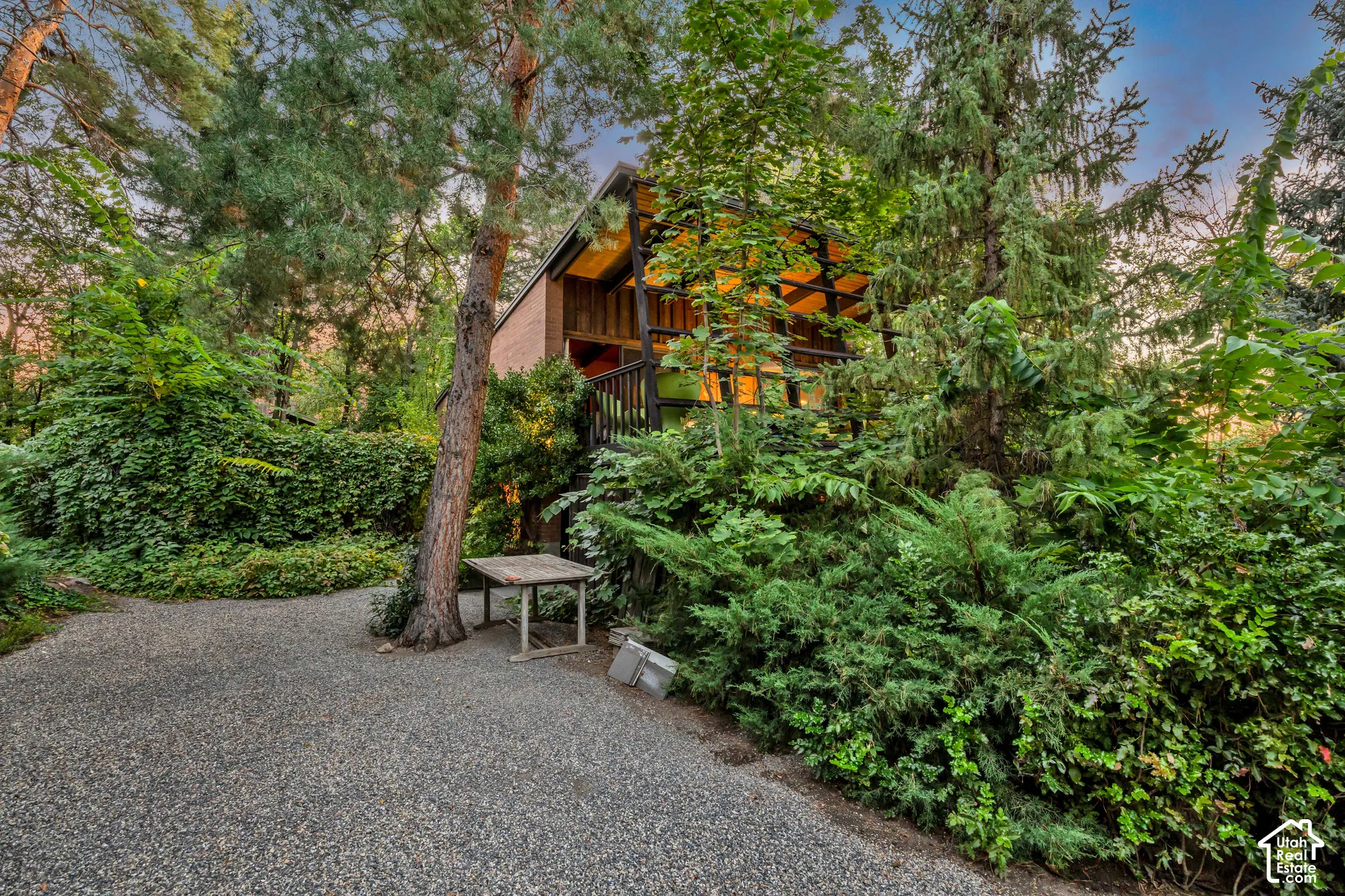 Gravel driveway leading to a residence featuring a wood balcony and large, exposed wood-frame windows