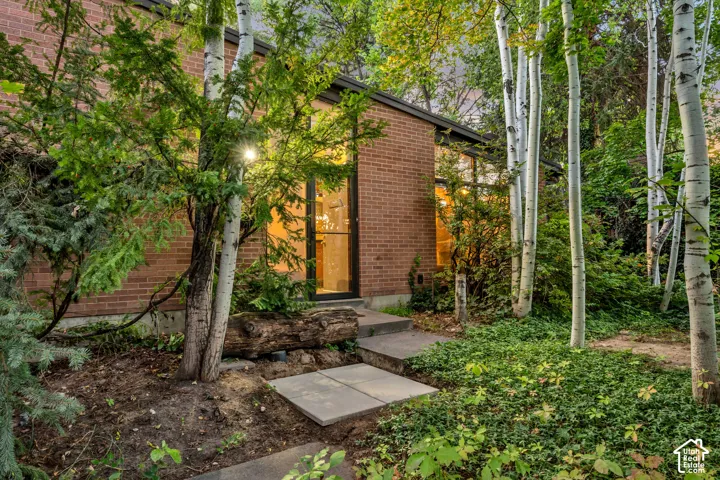 Brick exterior with a prominent full-height window, black-framed door, mature trees, and concrete stepping stones