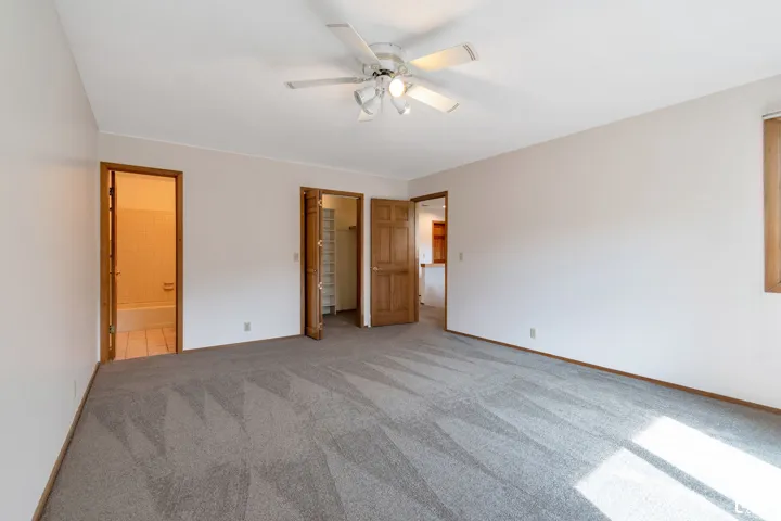 Spacious room featuring neutral carpeting, a ceiling fan with integrated lighting, and natural wood trim around doorways and the window