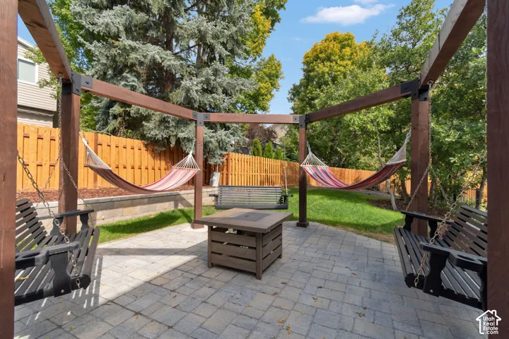 Expansive outdoor entertaining space featuring a wood pergola structure with integrated hammocks, paver patio flooring, a central fire pit with seating, mature landscaping with evergreen and deciduous trees, and yellow privacy fencing throughout the perim