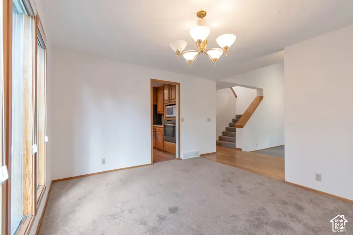 Spacious living area featuring hardwood flooring, abundant natural light from large windows, a brass and glass chandelier, and an open layout with access to a wooden staircase and closeted storage spaces