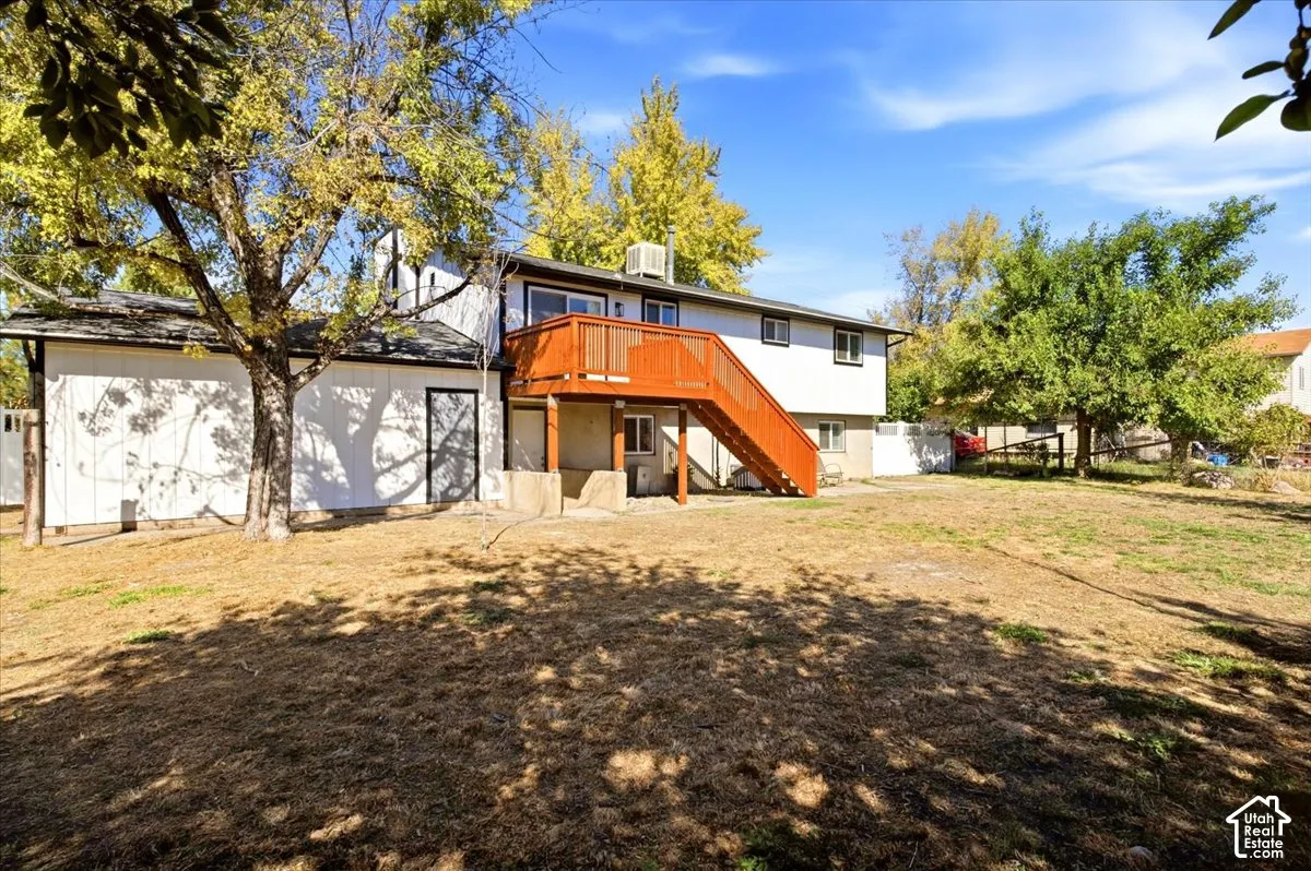 Expansive rear yard with mature trees, a two-story residence featuring white siding, a prominent wood-finish deck with stairs, and multiple windows