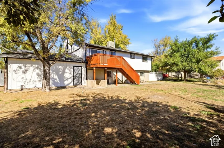 Expansive rear yard with mature trees, a two-story residence featuring white siding, a prominent wood-finish deck with stairs, and multiple windows