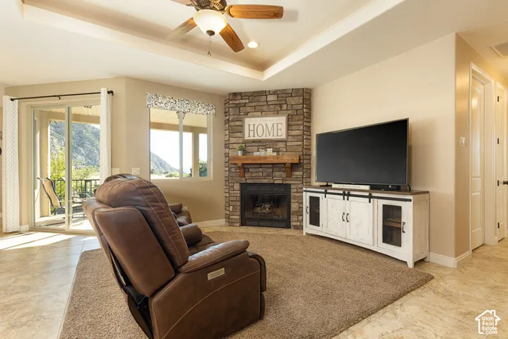Corner stone-clad fireplace with wood mantle, tray ceiling with a built-in ceiling fan, glass sliding door leading to an outdoor area, window with a valance, and tile flooring