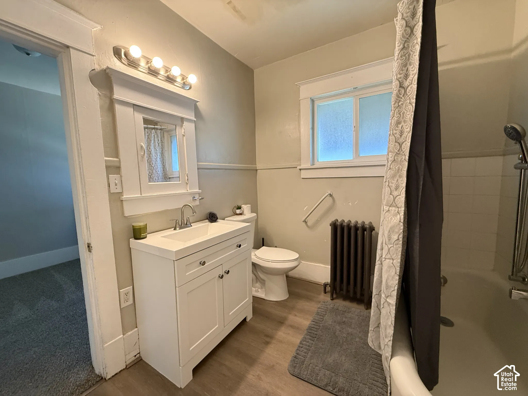 Bathroom featuring a white vanity cabinet with a rectangular basin, mirrored medicine cabinet, metal radiator, sliding window, and wood-finish flooring