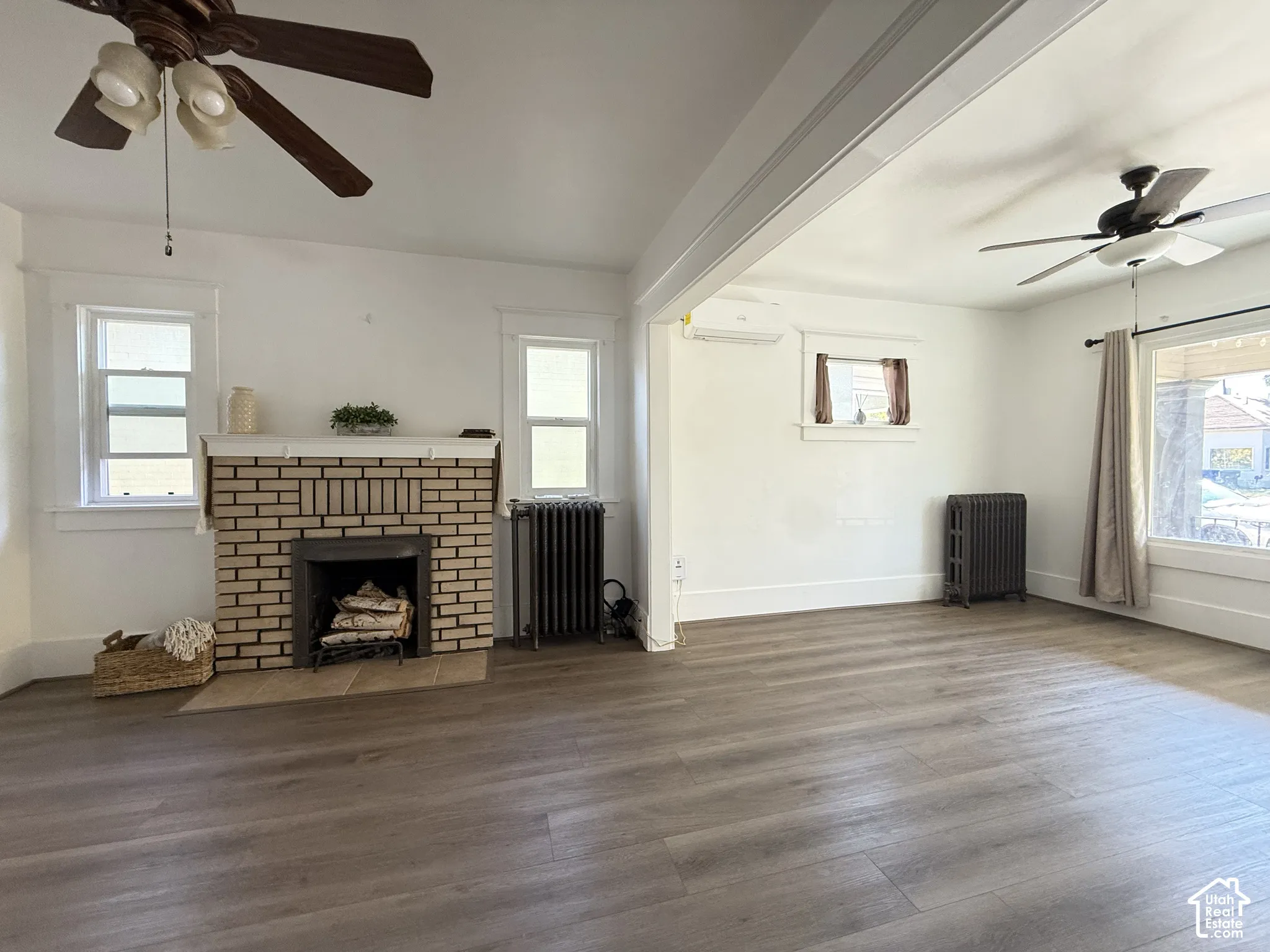 Living area featuring wood-finish flooring, a brick fireplace with a mantle, dual ceiling fans, wall-mounted ductless air conditioning, and cast-iron radiators
