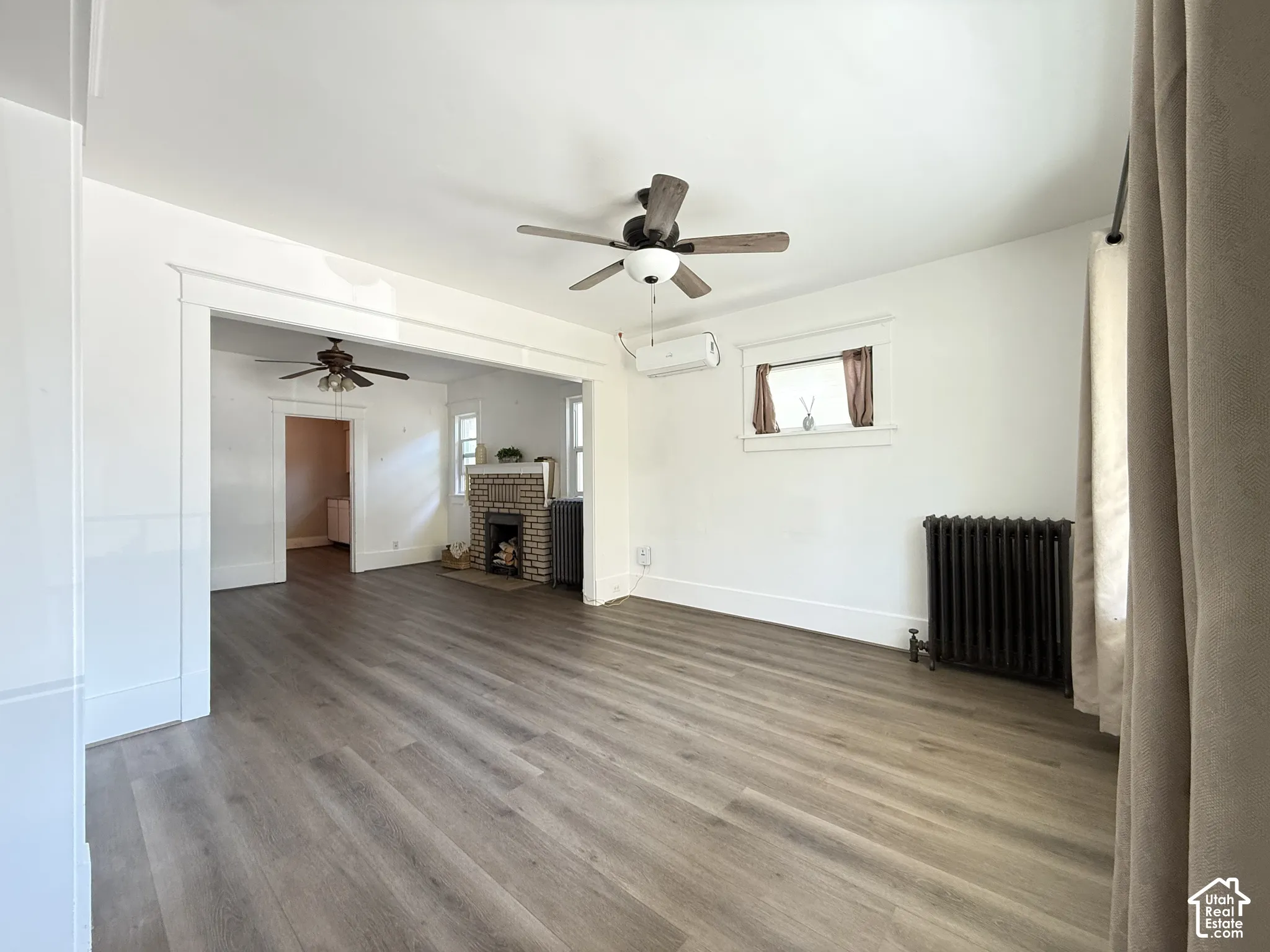 Wood-finish flooring throughout featuring a brick-surround fireplace, ceiling fans, wall-mounted heating and cooling unit, and cast iron radiator