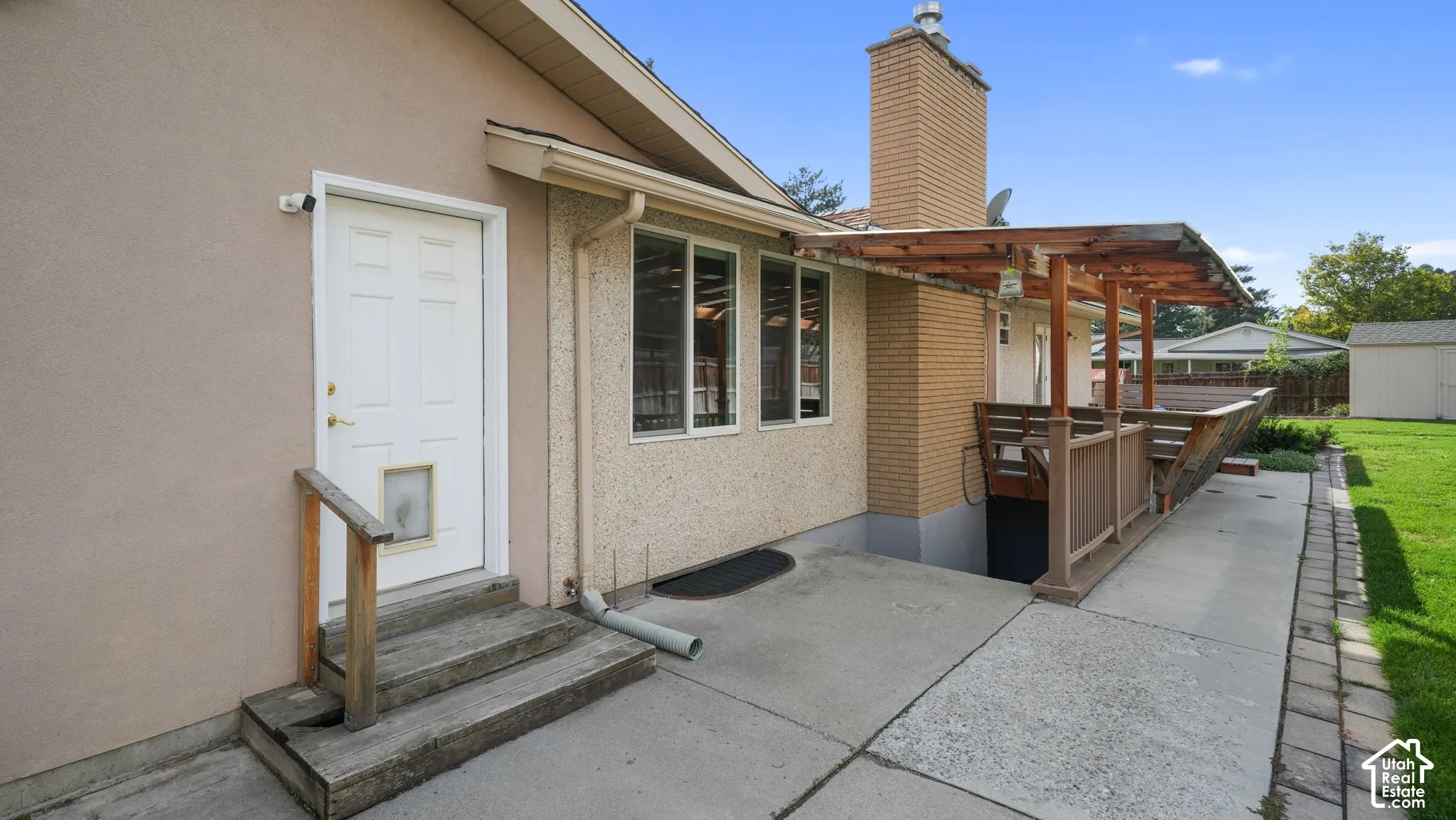 Exterior features a stucco finish, multi-pane windows, a wooden deck with a pergola, a brick chimney, and a concrete patio with a paver border