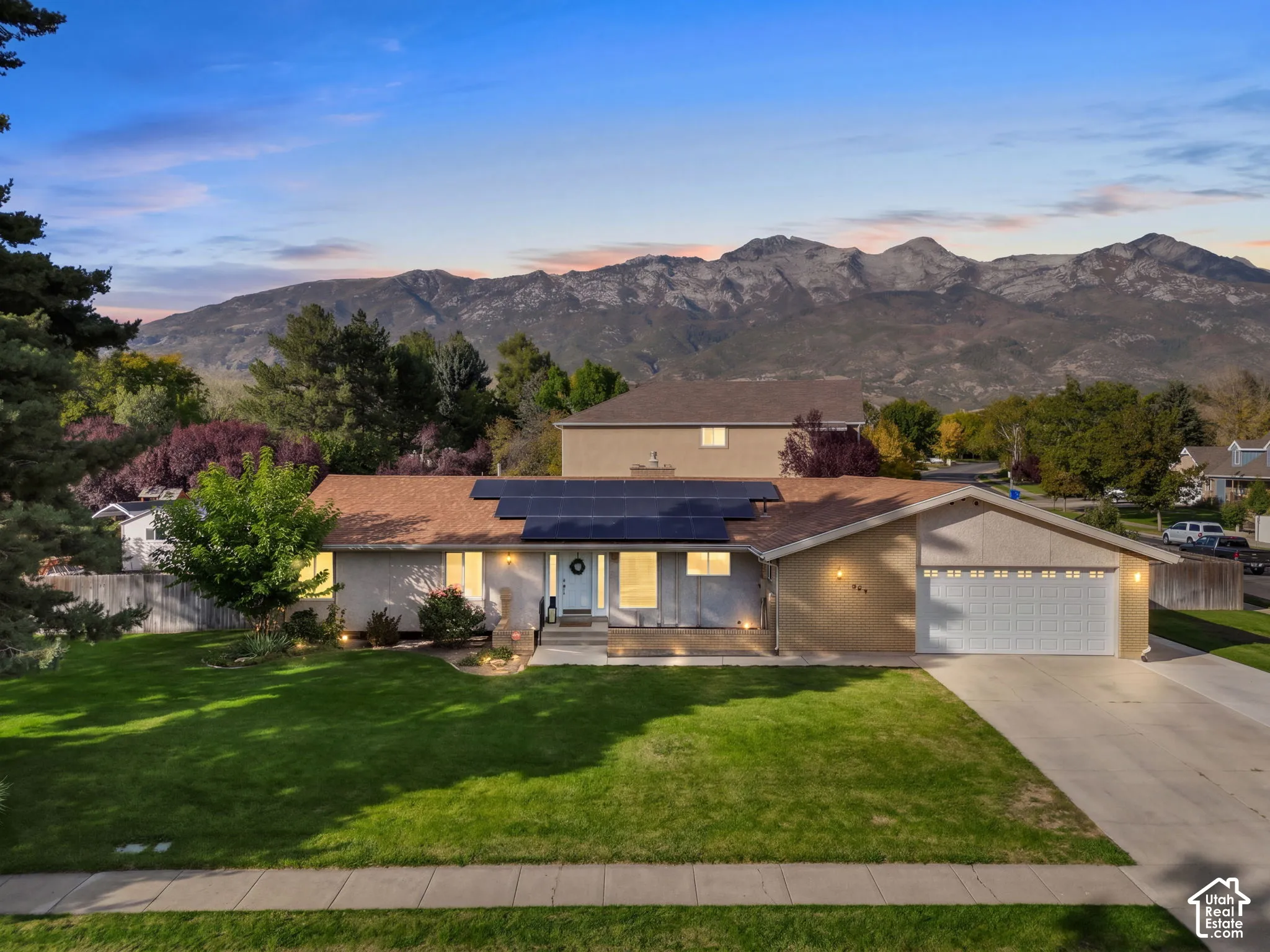 Expansive mountain backdrop, front yard with mature trees, established landscaping, solar panels, and an attached two-car garage