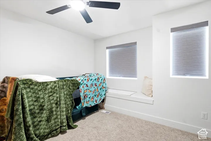 Plush carpet flooring, integrated ceiling fan with lighting, two windows with roller shades, and a built-in window seat