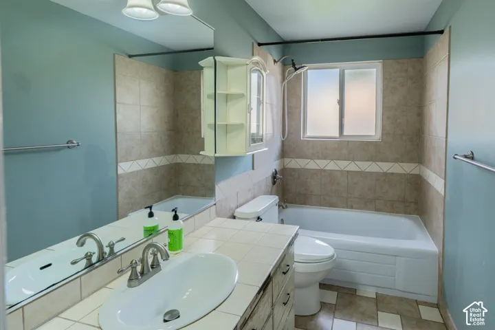 Vanity with ceramic tile countertop and integrated sink, full-width mirror, tile flooring, and tiled shower/tub combination with window