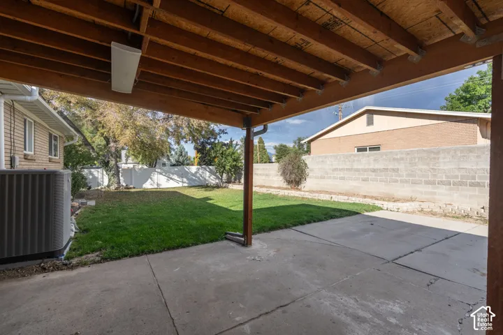 Covered patio with concrete flooring, exposed wood beam ceiling, and a large grassy yard featuring a white privacy fence