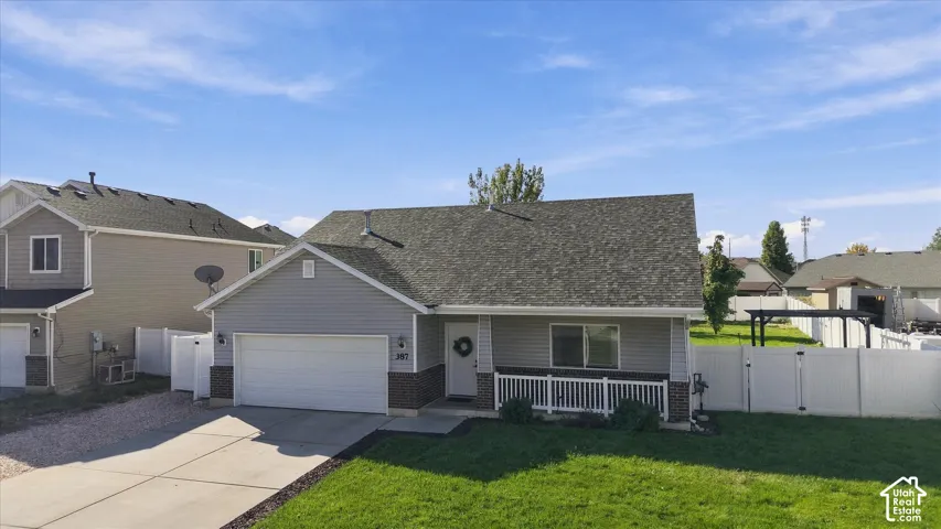 Light gray siding with dark brick accent, attached two-car garage, white front porch railing, green lawn, and a fenced backyard