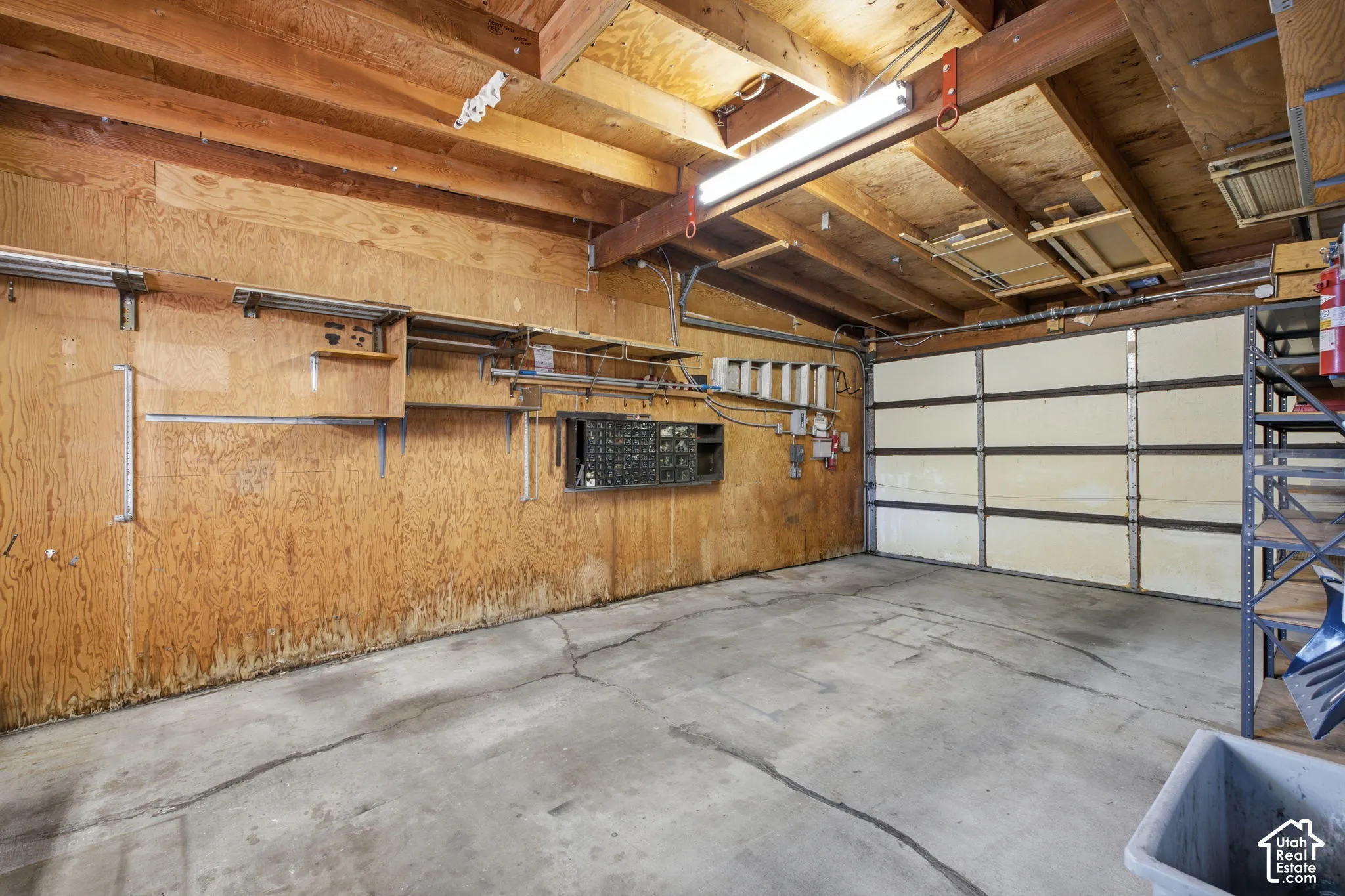 Concrete floor, exposed wood beam ceiling, plywood wall paneling, fluorescent lighting fixture, overhead garage door