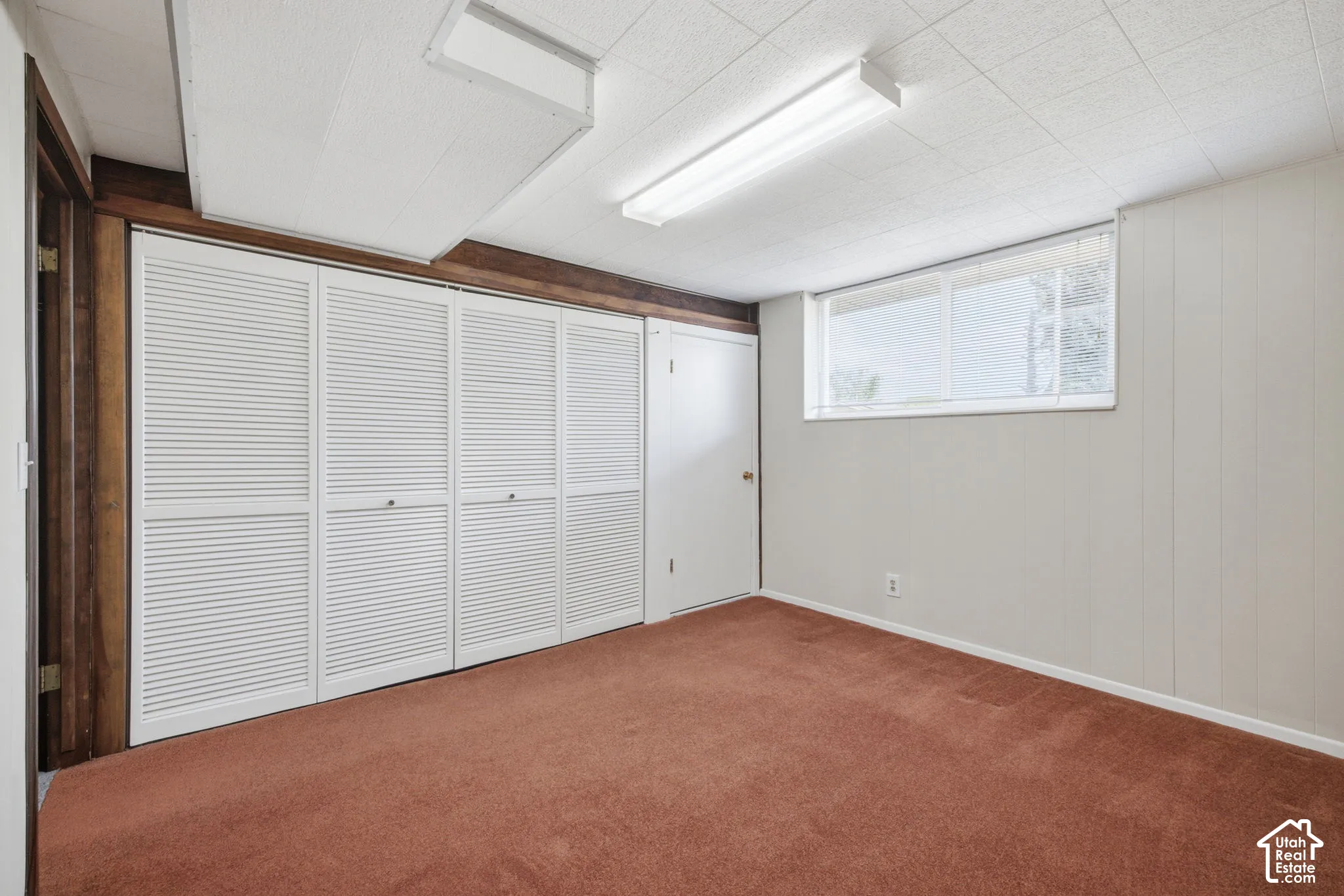 Textured acoustic ceiling tiles with fluorescent lighting, louvered bi-fold closet doors, wood paneling, and a single window with blinds