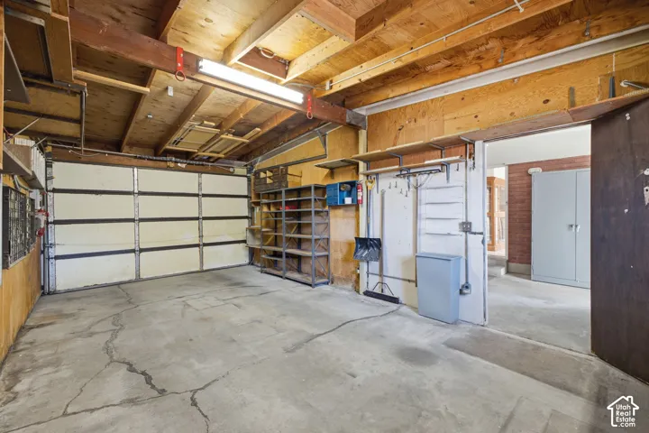 Concrete flooring with an overhead fluorescent light fixture, exposed wood beam ceiling, and a paneled garage door