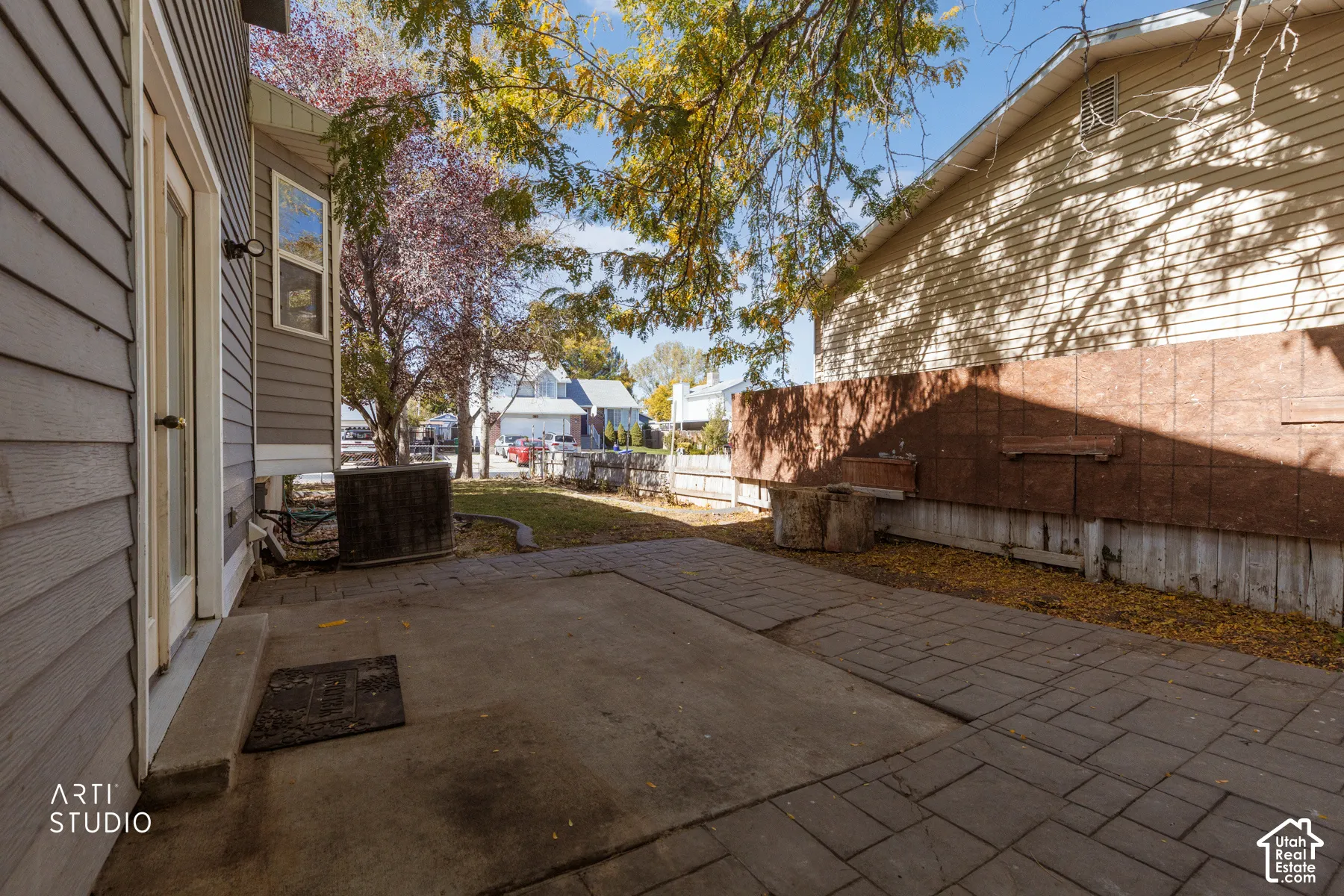 Paved patio area, gray siding exterior, rear entry door with glass panel, mature trees, and landscaping
