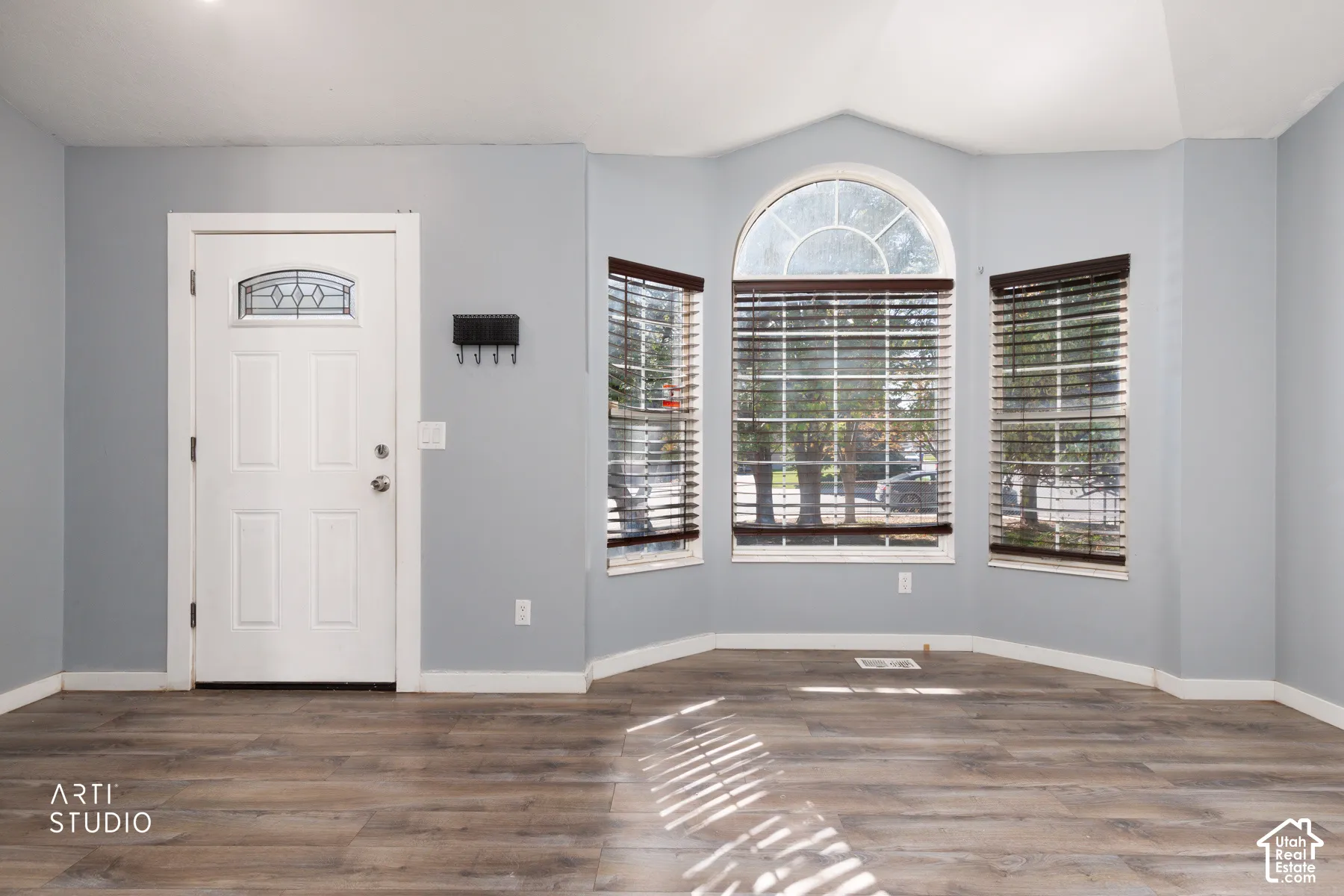 Wood-finish flooring throughout, bay windows with blinds, arched transom window, and a paneled entry door with decorative glass insert
