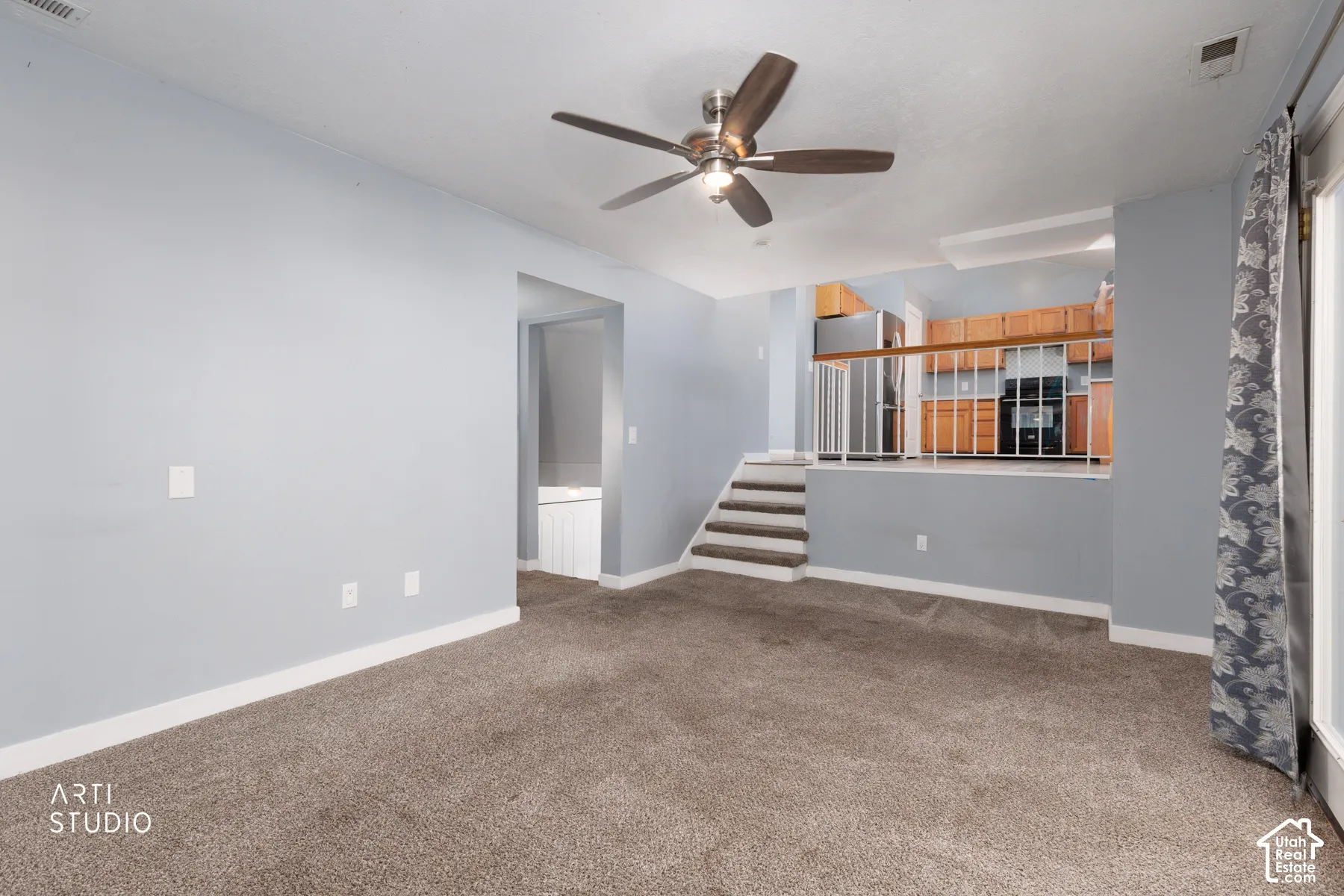 Carpeted living area featuring a ceiling fan, recessed kitchen with wood-finish cabinetry, and a carpeted staircase with a white railing