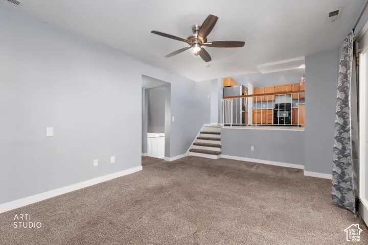 Carpeted living area featuring a ceiling fan, recessed kitchen with wood-finish cabinetry, and a carpeted staircase with a white railing