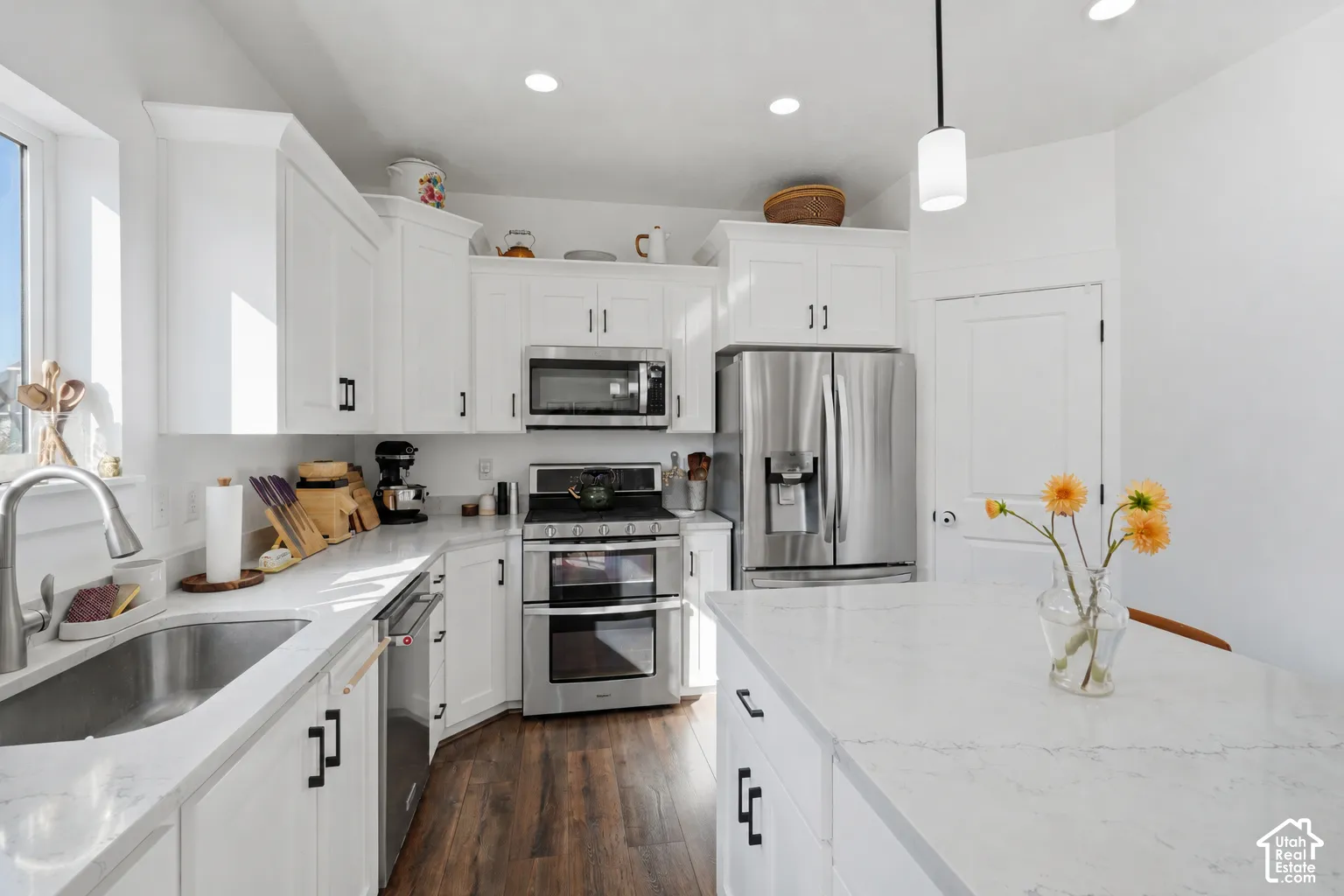 White cabinetry with black hardware, stainless steel appliances, wood-finish flooring, recessed lighting, and a kitchen island with a light-toned countertop