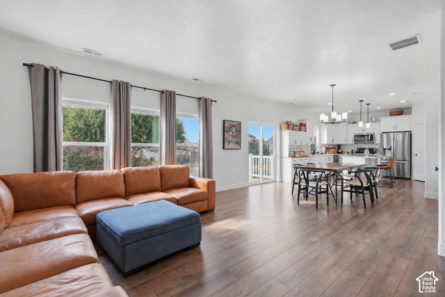 Spacious open-concept living area featuring wood-finish flooring, a kitchen with white cabinetry, stainless steel appliances, and a dining area with pendant lighting