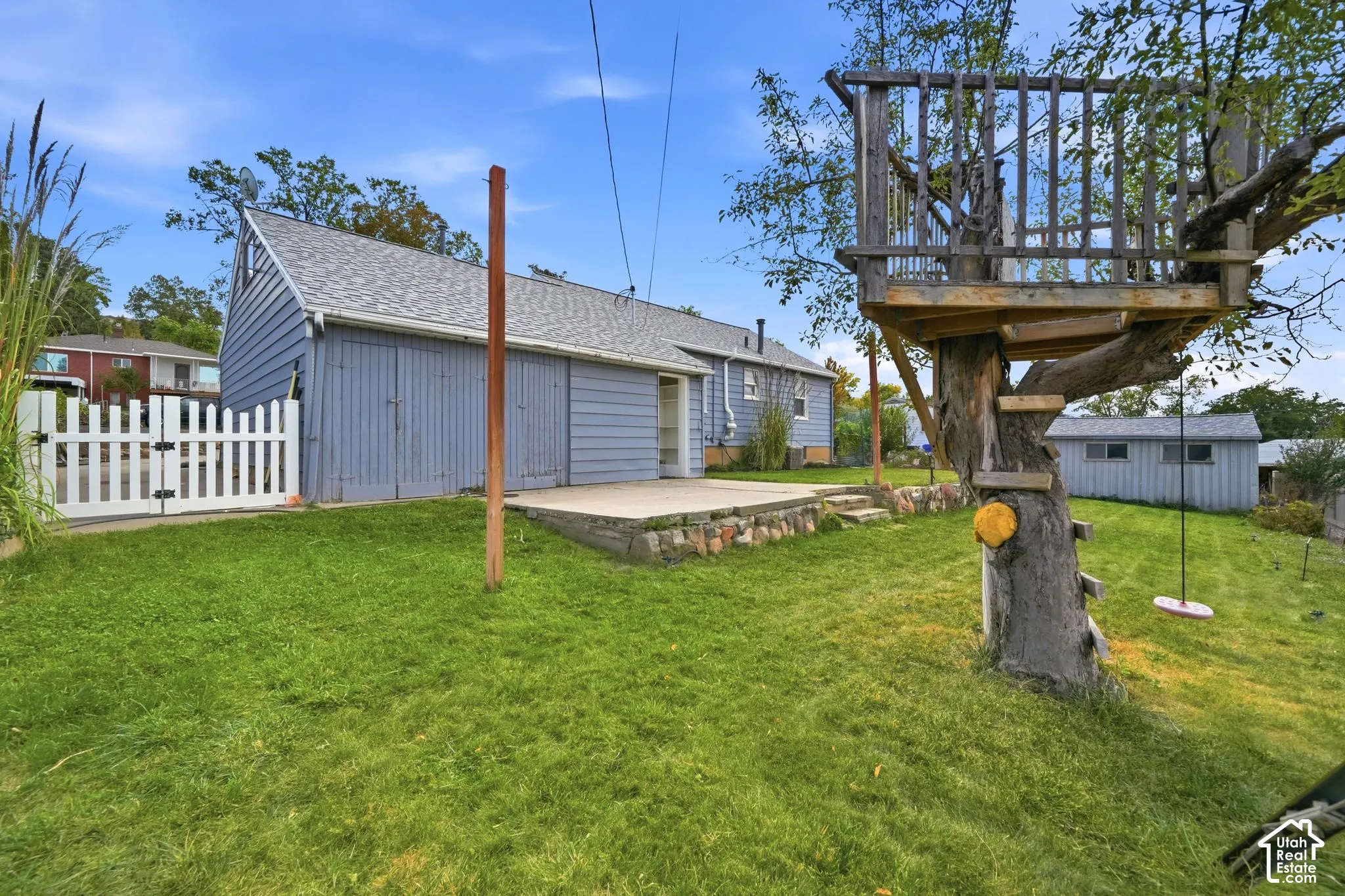 Expansive green lawn, white picket fence, concrete patio, detached storage structure, and mature tree with built-in platform