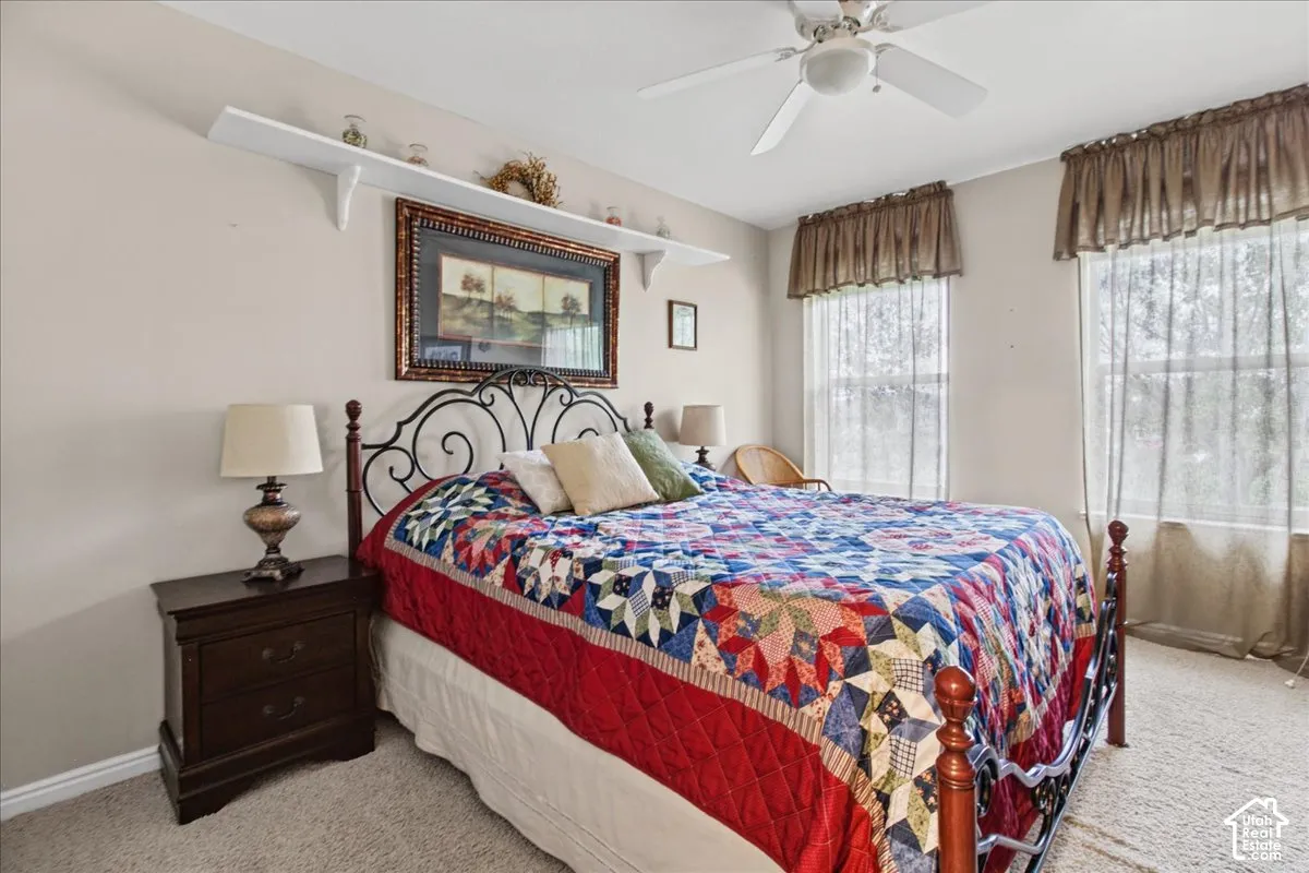 Two windows with textile window treatments, ceiling fan with light fixture, wall-mounted shelving, and carpeted flooring