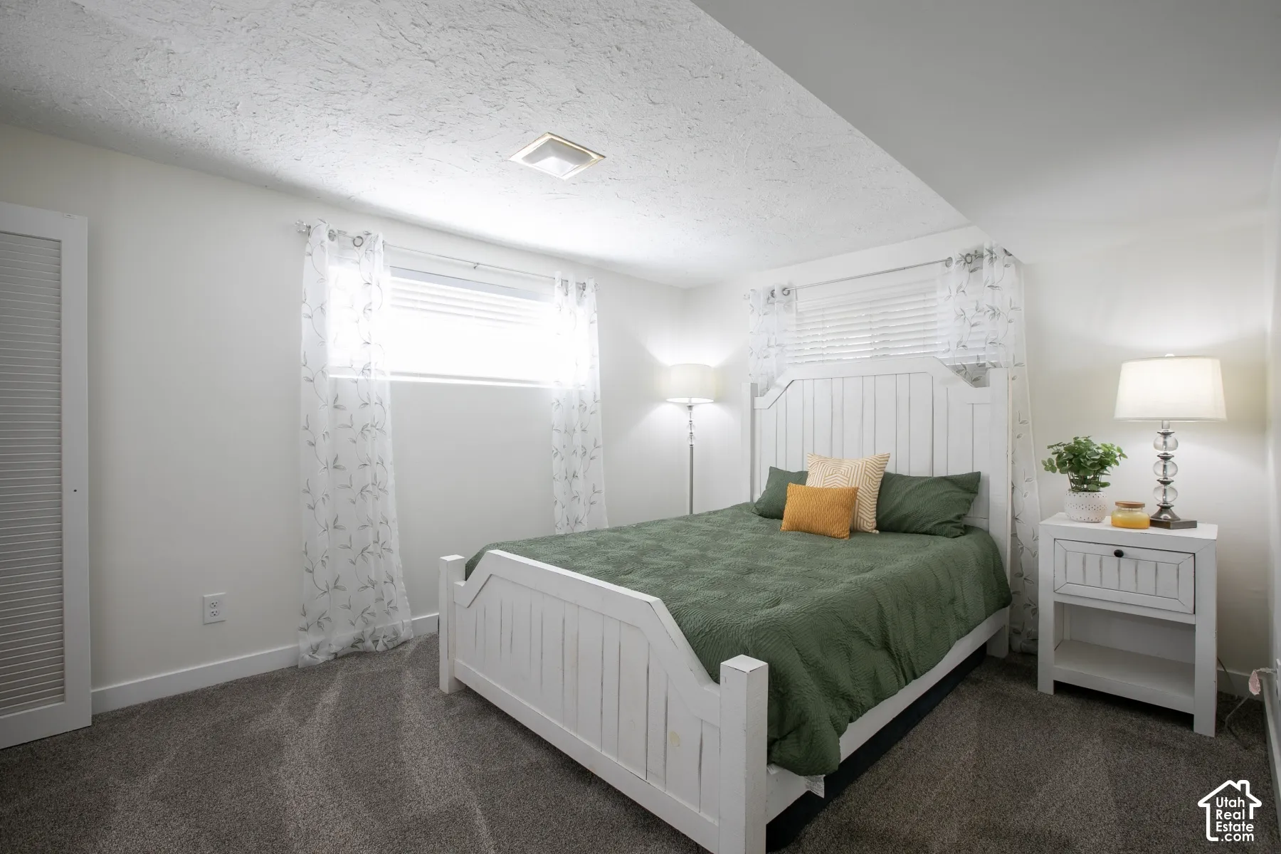 White textured ceiling, gray carpet flooring, window with blinds, and a white paneled bed frame