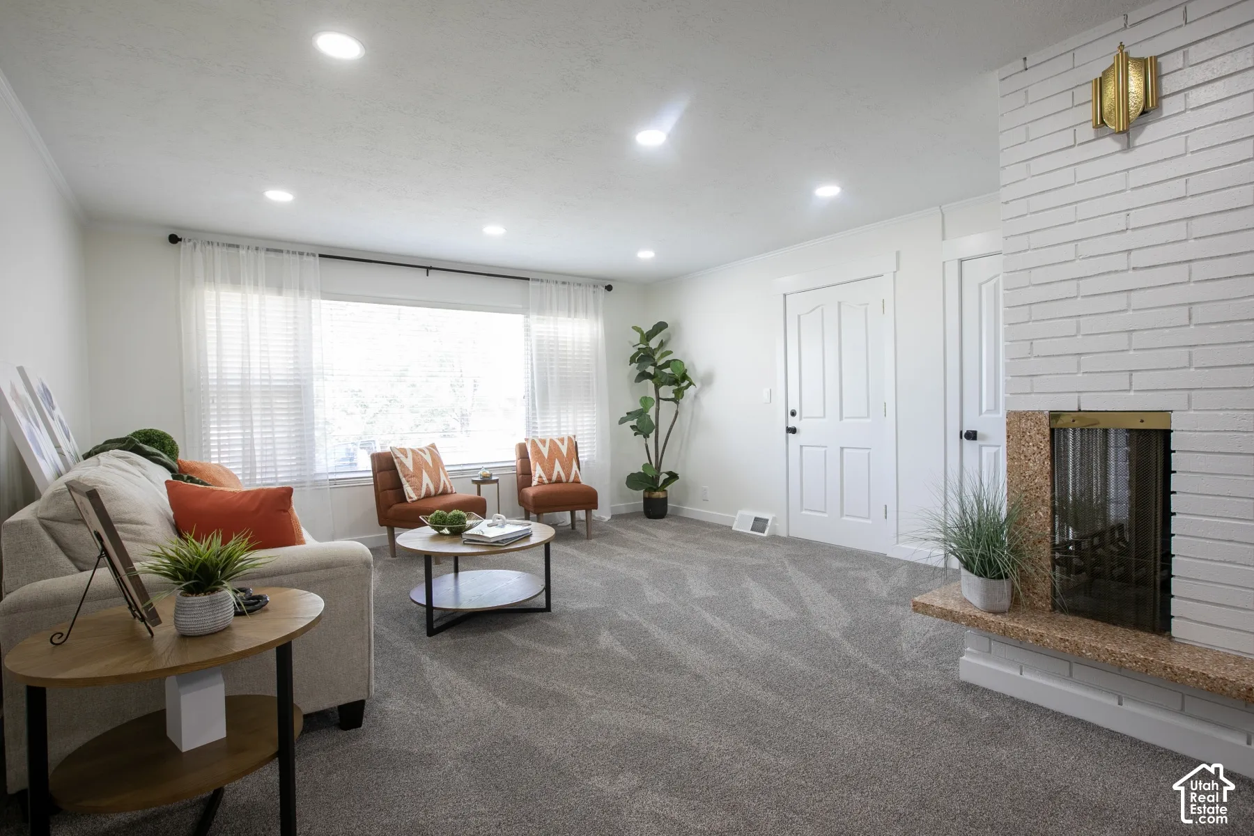 White painted brick fireplace with a stone hearth, recessed ceiling lighting, a large window, and light gray patterned carpeting
