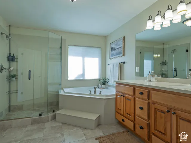 Large tiled flooring, glass-enclosed shower with showerhead, corner soaking tub, double vanity with light wood cabinetry, and a wide mirror