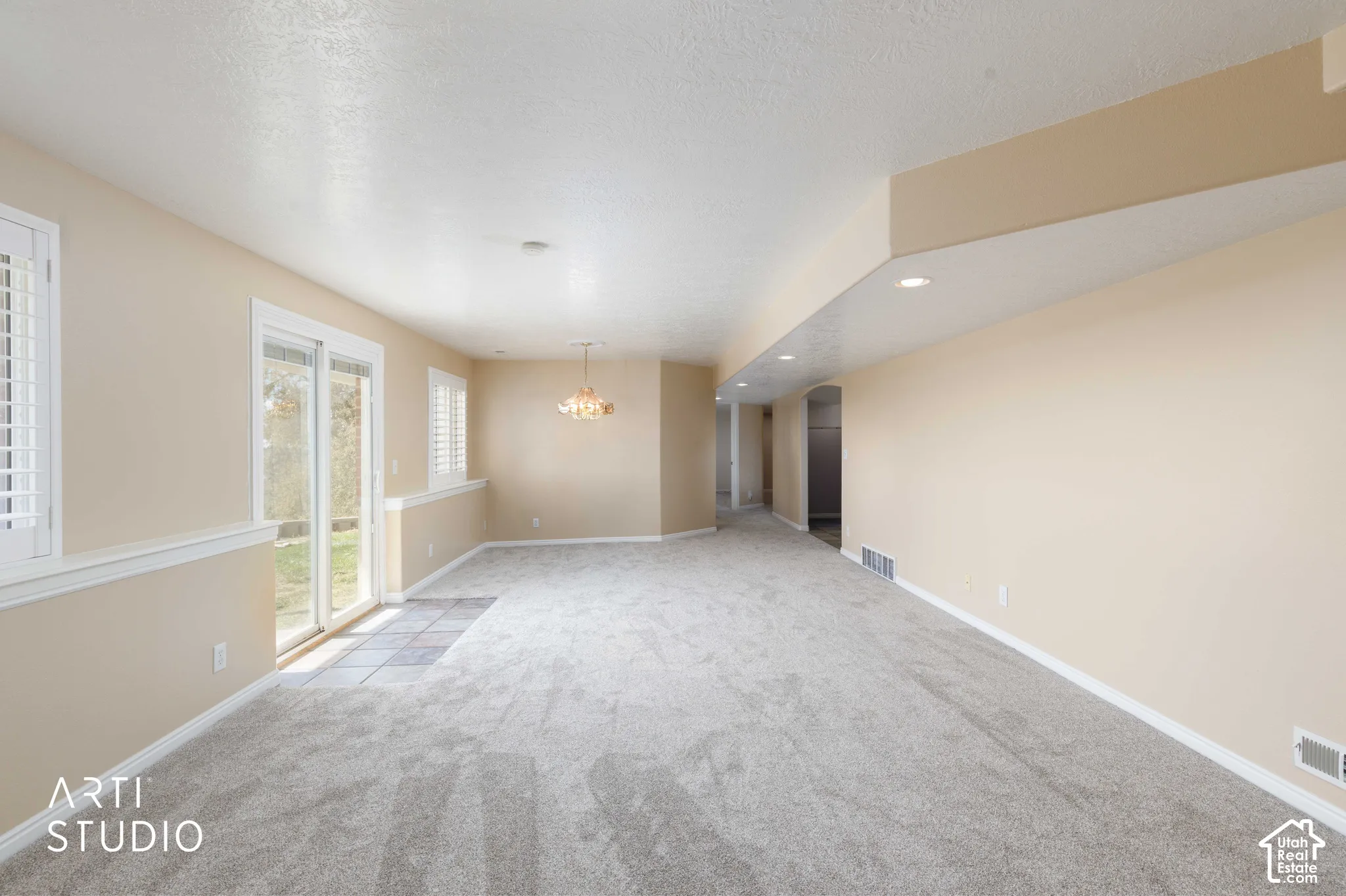 Spacious room featuring light-toned carpeting, sliding glass door, white plantation shutters, recessed lighting, and a decorative chandelier