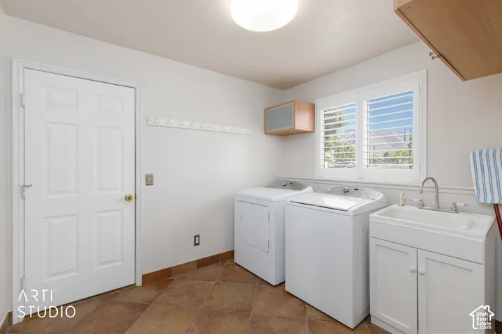 Laundry room featuring natural stone tile flooring, a utility sink with white cabinetry, a window with louvered shutters, and overhead cabinetry