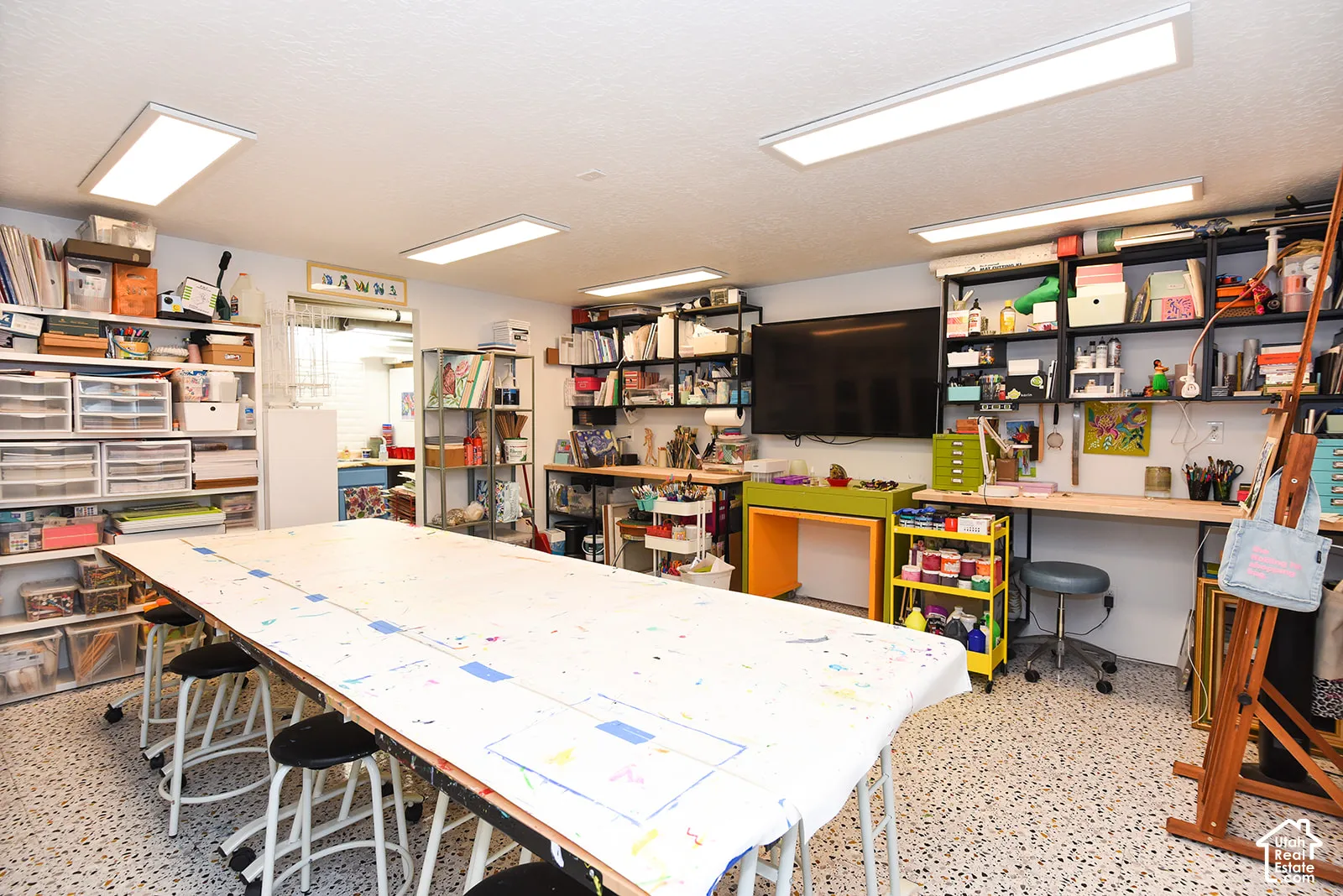 Dedicated studio workspace featuring expansive work surfaces, abundant natural lighting from recessed ceiling fixtures, extensive built-in shelving and storage throughout, concrete flooring, and a well-equipped layout designed for creative projects and or