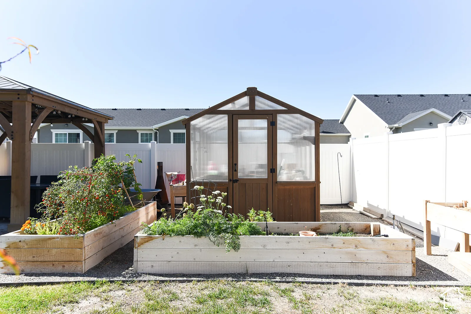 Backyard featuring a wooden gazebo, a brown greenhouse structure, two raised garden beds, and white privacy fencing