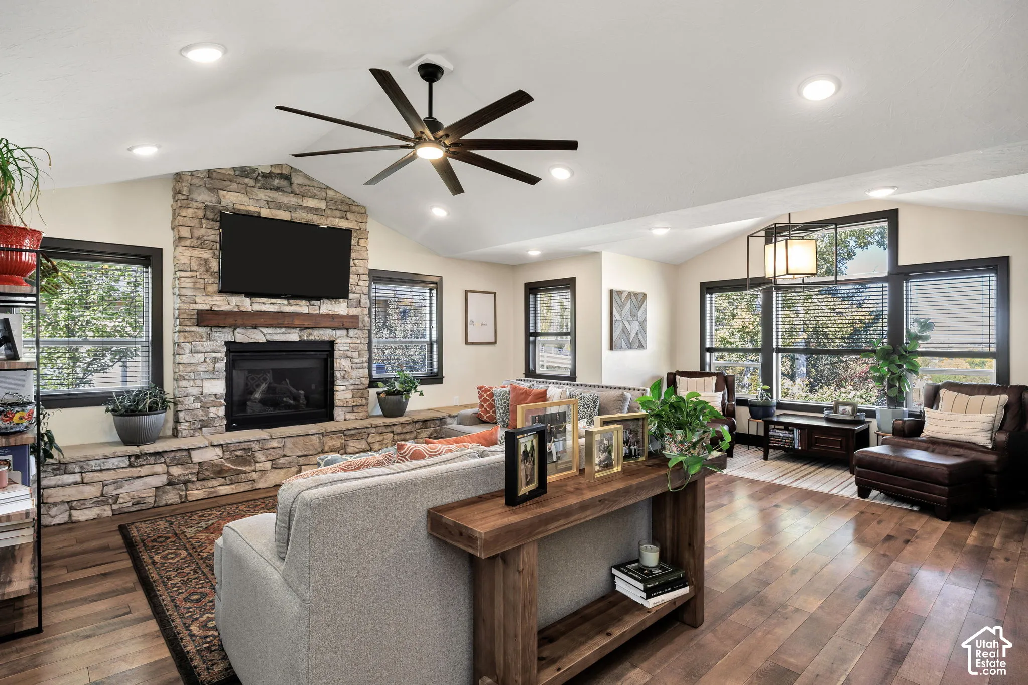 Vaulted ceiling with recessed lighting, wood-finish flooring, stone-faced fireplace with wood mantel, large windows with dark trim, and a ceiling fan