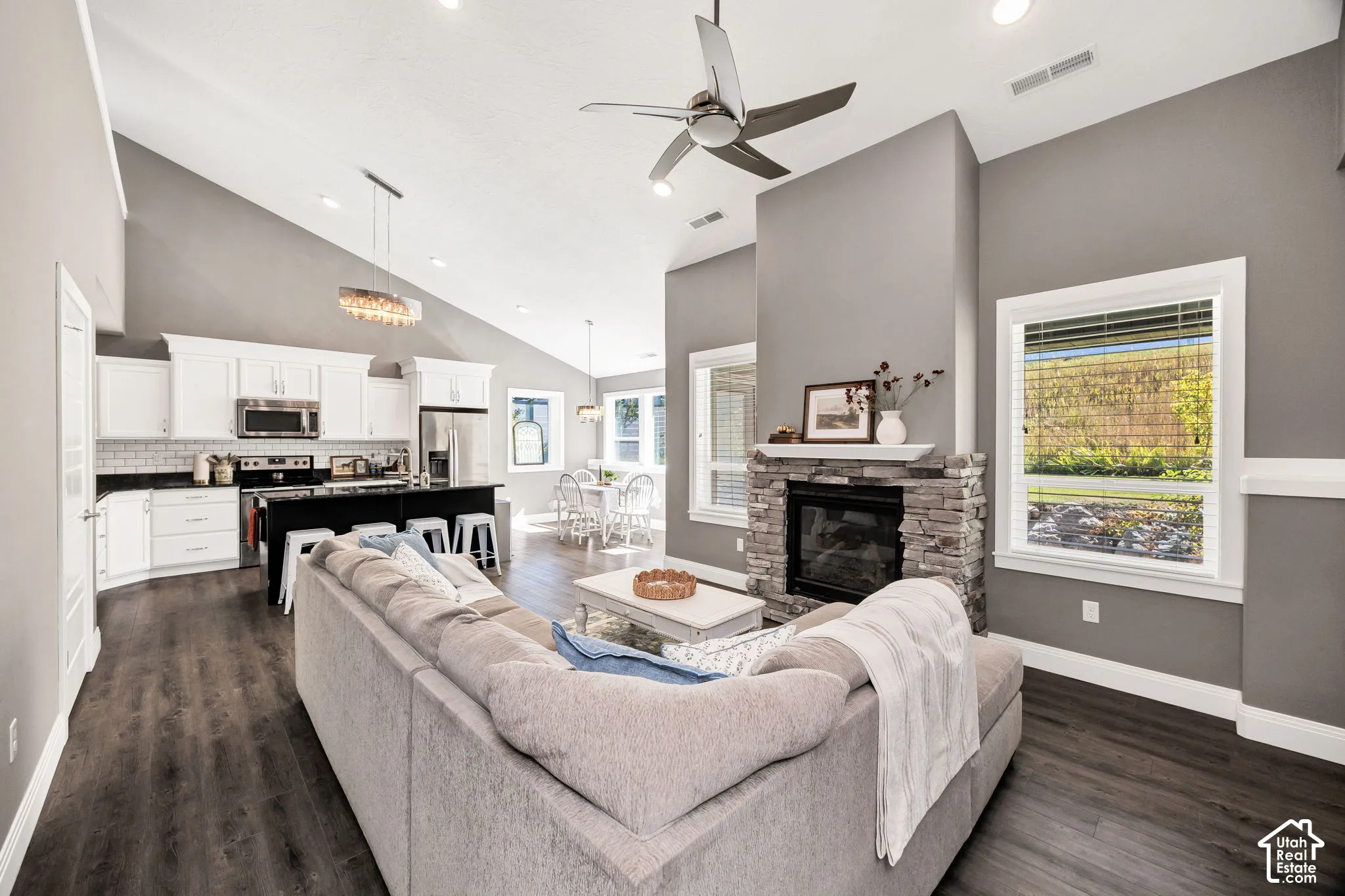 Open-concept living area featuring vaulted ceilings, wood-finish flooring, a stone-clad fireplace, white cabinetry, and a kitchen island with seating