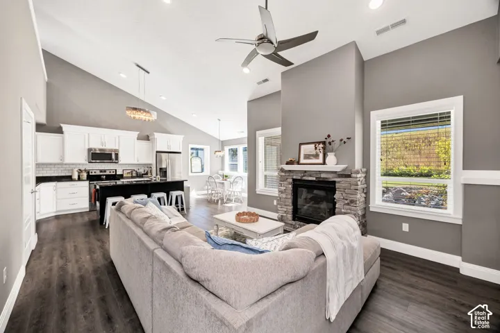 Open-concept living area featuring vaulted ceilings, wood-finish flooring, a stone-clad fireplace, white cabinetry, and a kitchen island with seating
