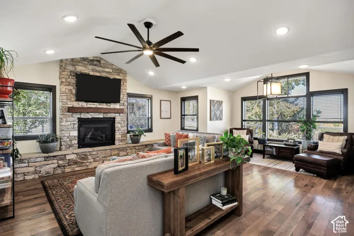 Vaulted ceiling with recessed lighting, wood-finish flooring, stone-faced fireplace with wood mantel, large windows with dark trim, and a ceiling fan