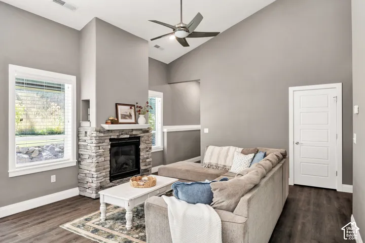 Vaulted ceiling with a ceiling fan, stone-clad fireplace with a mantel, wood-finish flooring, and white trim windows
