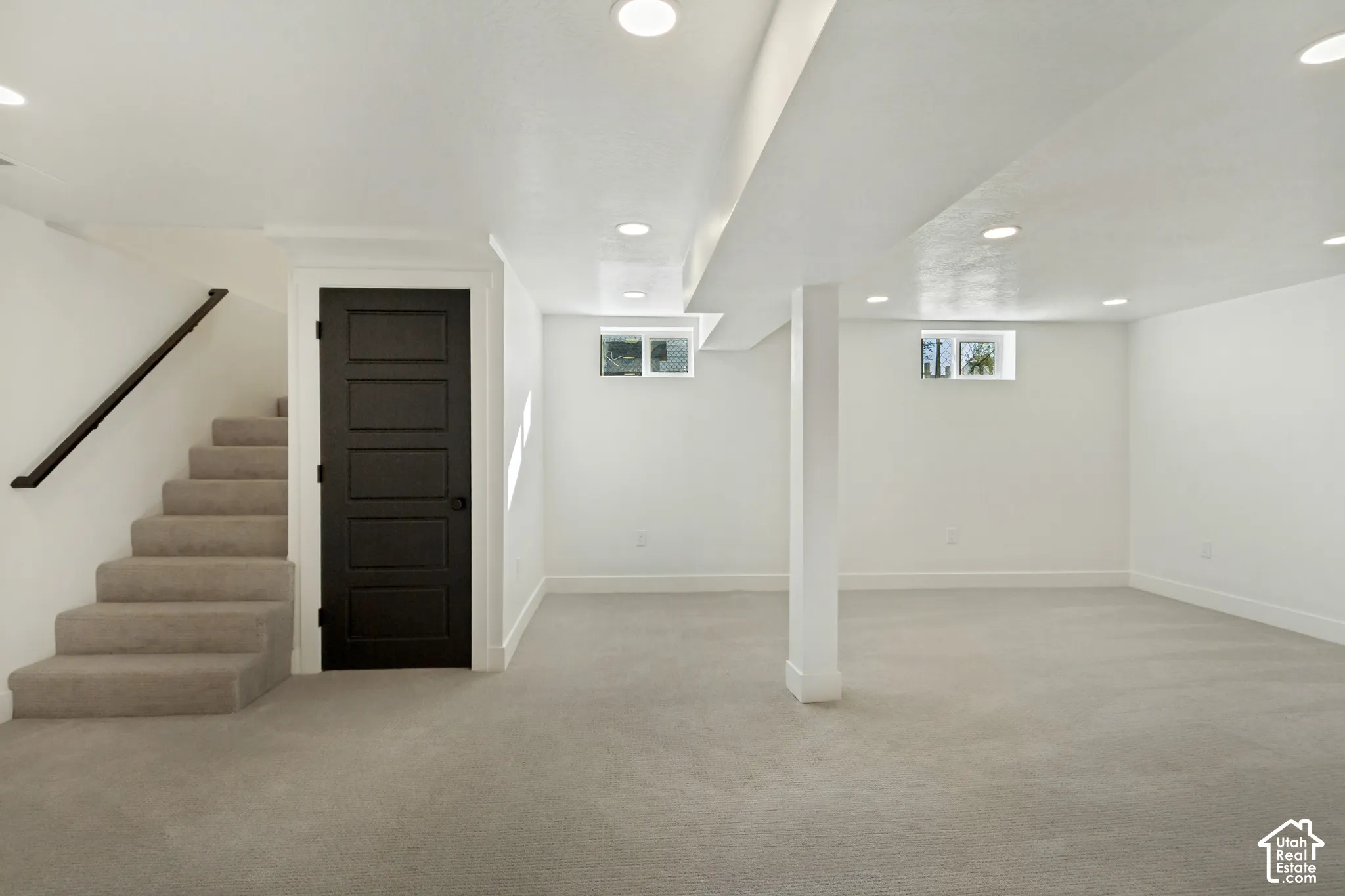 Expansive carpeted area with recessed lighting, a fully finished staircase with a dark railing, a five-panel interior door, and permanent windows