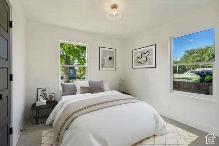 Serene bedroom featuring abundant natural light from dual large windows, neutral white walls, light wood-finish flooring, and a geometric patterned area rug