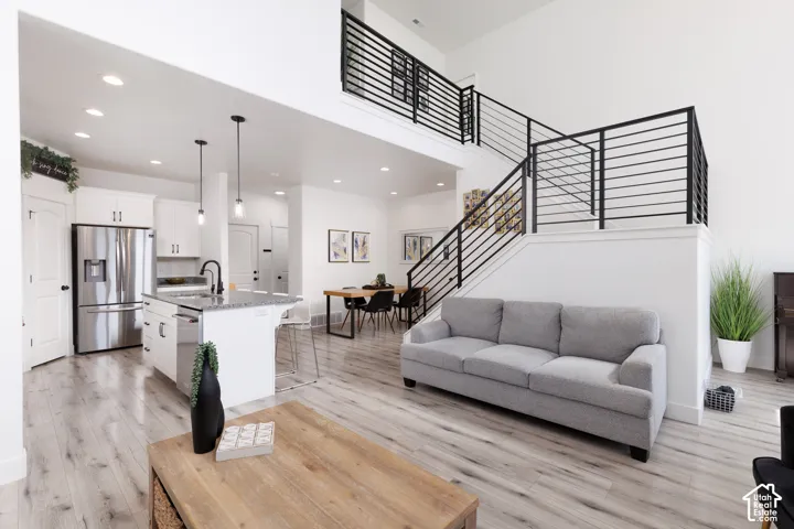Open-concept living area with high ceilings, wood-finish flooring, a kitchen island with seating, white cabinetry, and a modern metal staircase railing