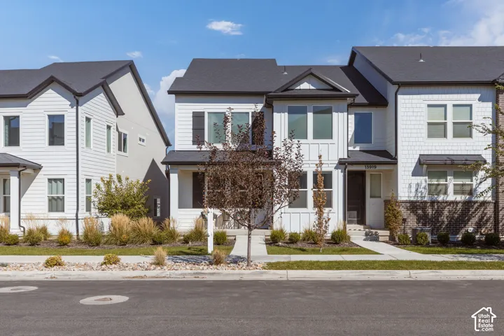 Contemporary townhome featuring white siding, dark trim, a dark asphalt shingle roof, and a mature tree